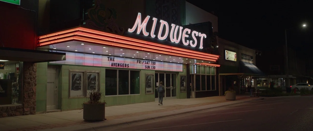 Nomadland (2020) movie still: wide — A small town cinema at night with green tiled walls and a bright neon sign. A lone person with a backpack walks…