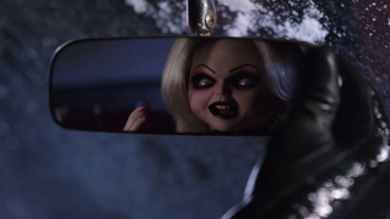 Cult of Chucky (2017) movie still: closeup — The interior of a vehicle at night with a blurred, dark background visible. A doll with blonde hair and…