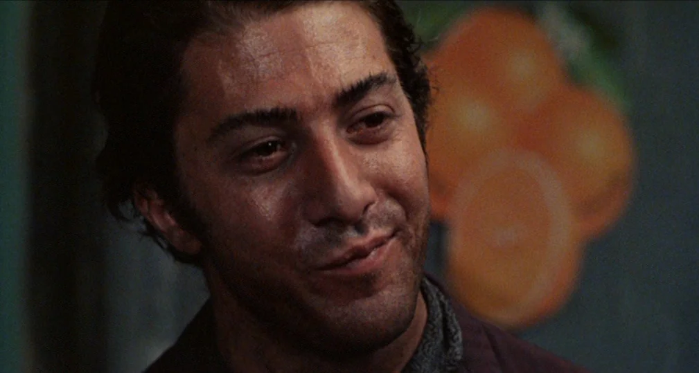 Midnight Cowboy (1969) movie still: closeup — A dimly lit indoor space features a wall advertisement with stylized orange imagery. A man with dark…