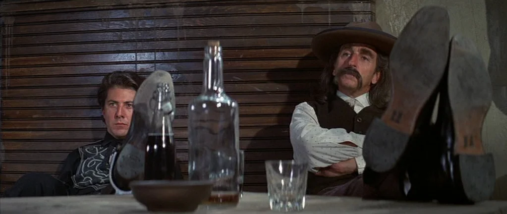 Little Big Man (1970) movie still: medium — A dimly lit room features a wooden wall and a table with bottles. Two men sit behind the table with…