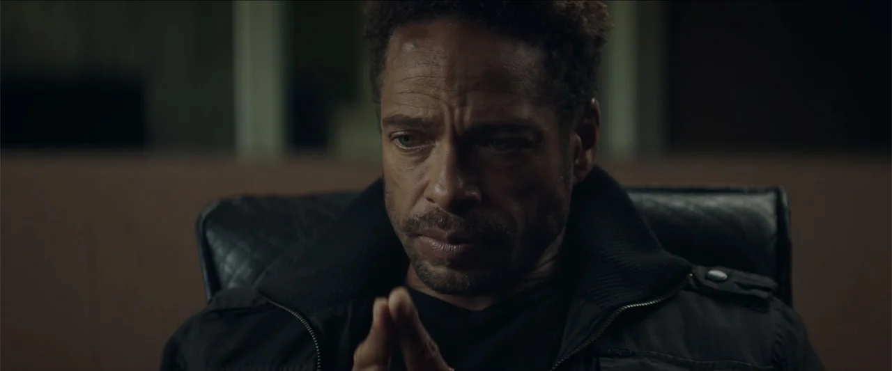 Redemption Day (2021) movie still: closeup — A dark, dimly lit office interior contains an out-of-focus leather chair in the background. A man sits…