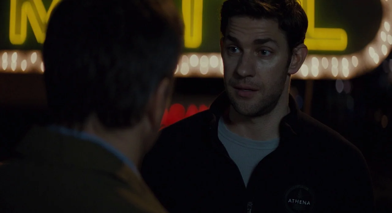 Promised Land (2012) movie still: medium — A man in a black fleece stands in a dark parking lot at night. An out-of-focus yellow neon motel sign is…