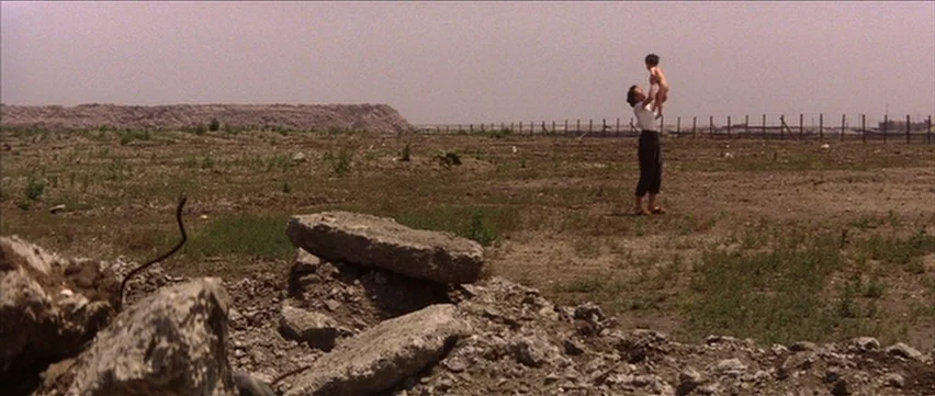 Gate of Flesh (1964) movie still: wide — A desolate and flat field with sparse vegetation extends toward a distant wire fence. A woman in light…