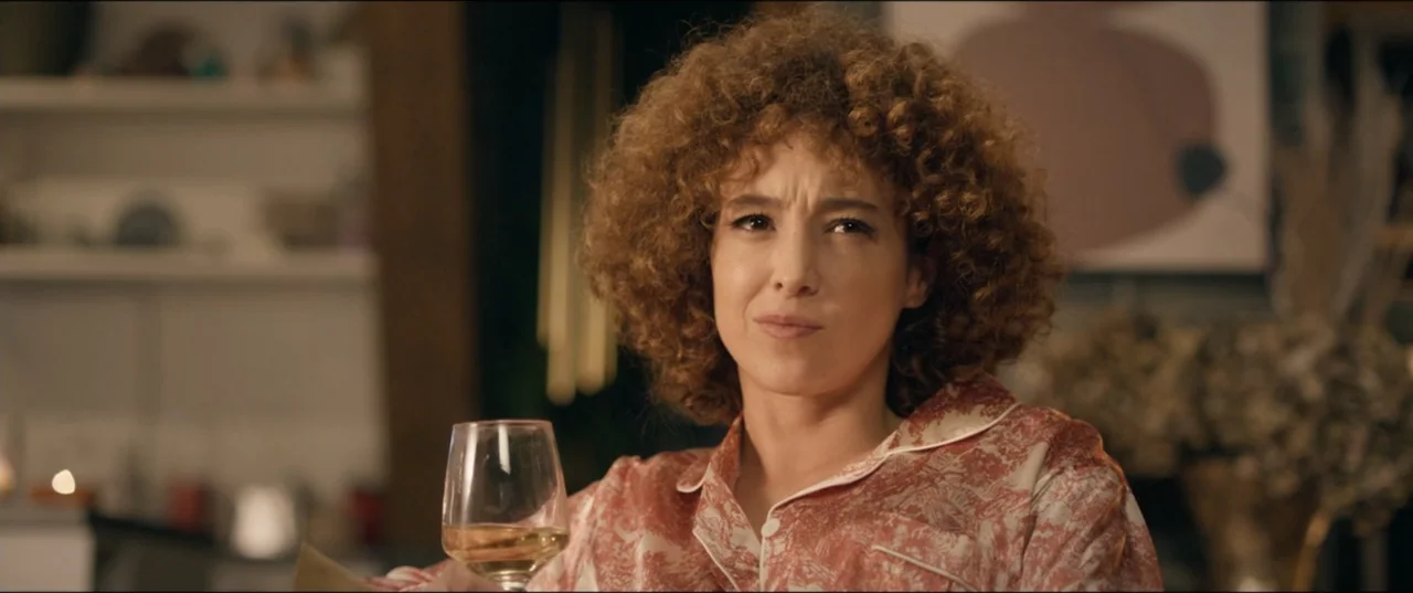 Risqué (2025) movie still: medium — A residential interior features a person sitting in a living room area. A woman wearing patterned pajamas holds…