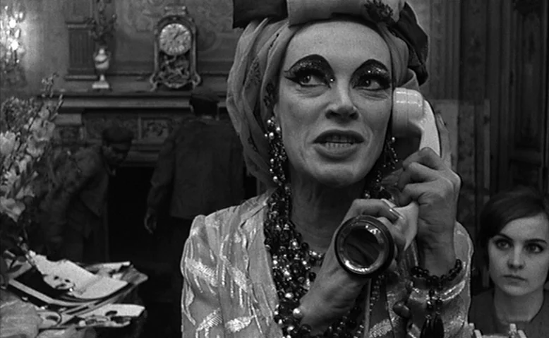 Who Are You, Polly Maggoo? (1966) movie still: closeup — An ornate room contains a fireplace with a mantel clock and floral arrangements. A woman…