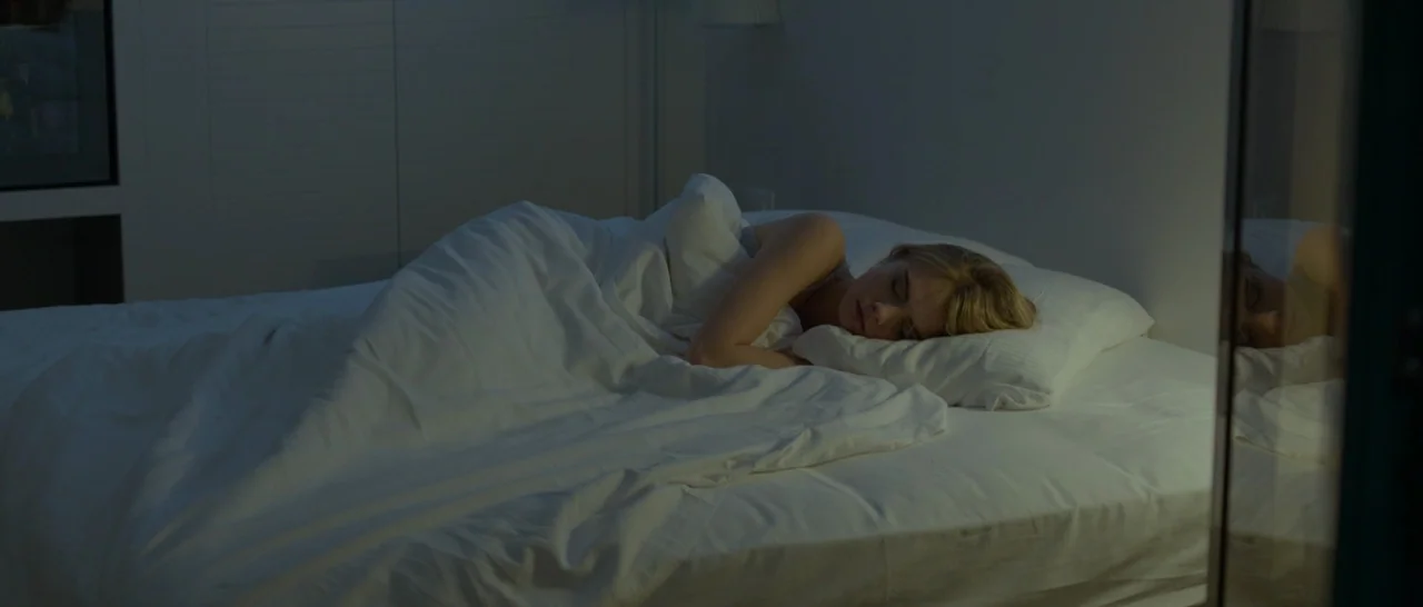 The Face of an Angel (2014) movie still: medium — A woman lies asleep in a bed with crisp white linens in a dim room. The subject is turned to her…