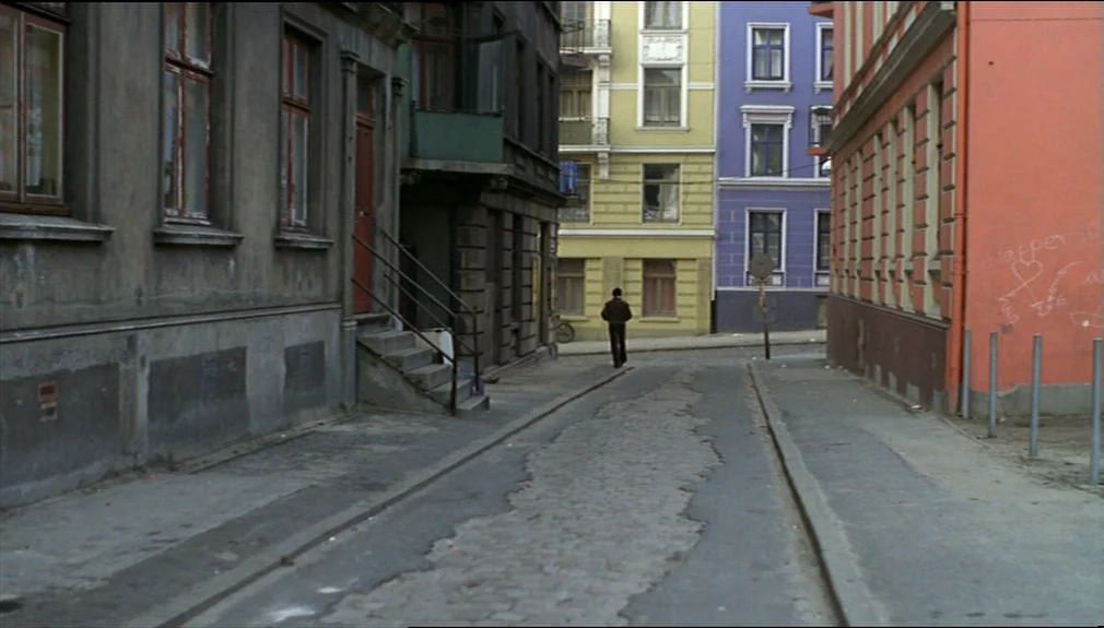 The American Friend (1977) movie still: wide — An urban intersection with aged multi-story tenement buildings painted in muted, contrasting colors.…