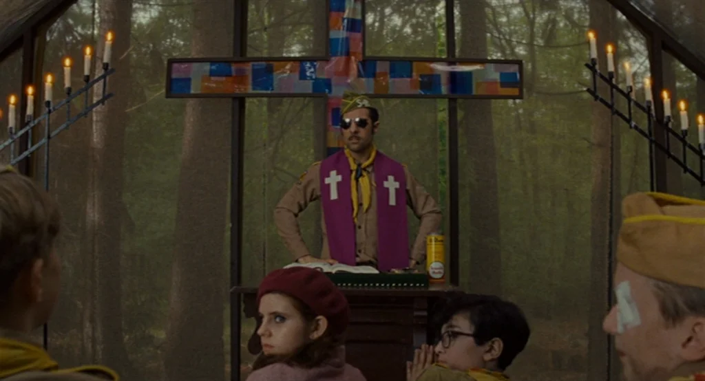 Moonrise Kingdom (2012) movie still: medium — A man in a scout uniform leads a service in an outdoor, woodland setting. A stained-glass cross is…