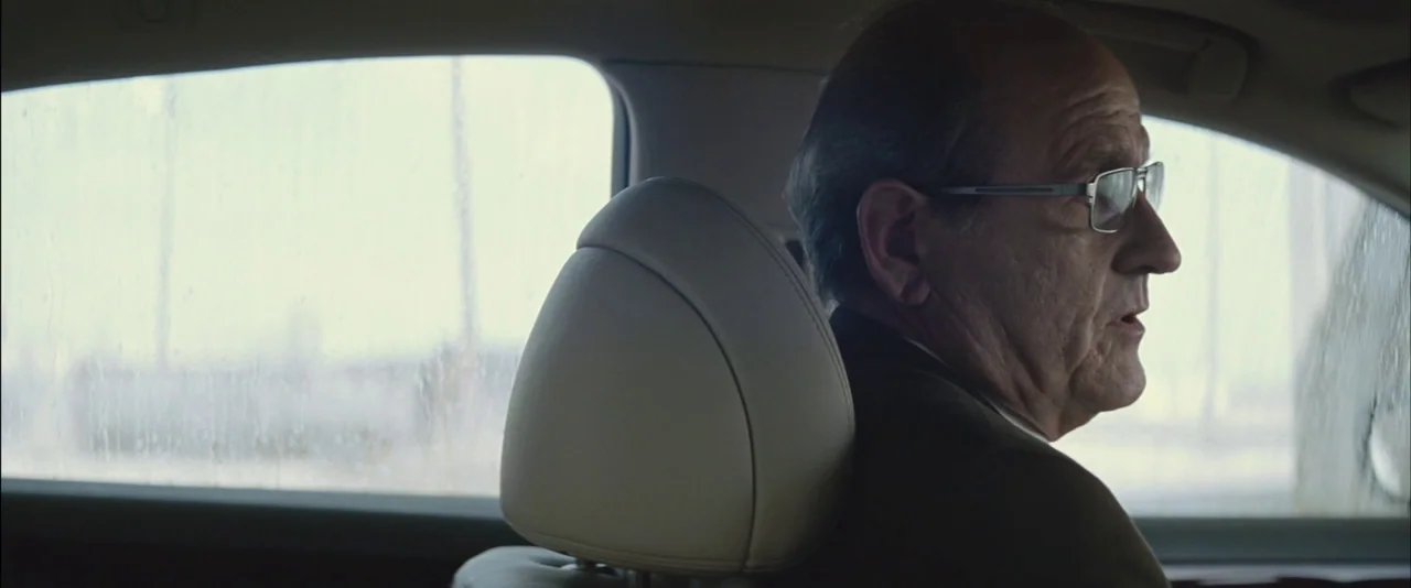 Killing Them Softly (2012) movie still: medium — The interior of a moving vehicle with rain streaks visible on the window glass. An elderly man in a…