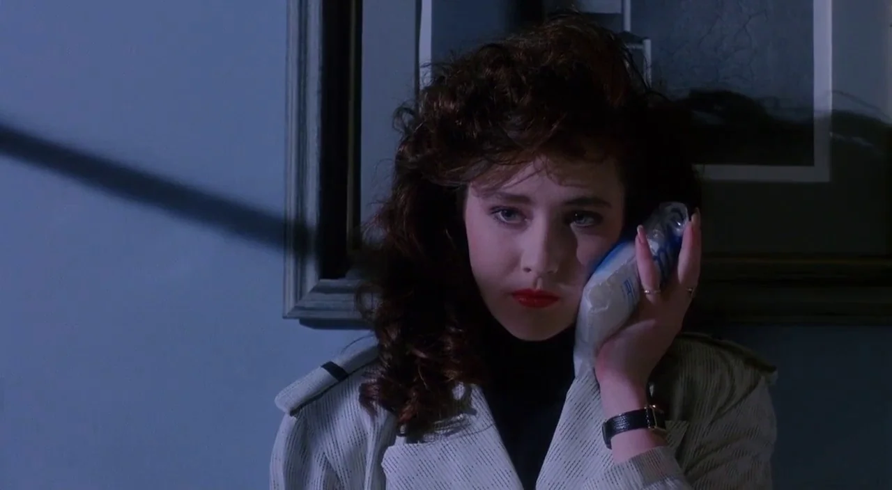 Heathers movie still: closeup — A dimly lit room interior features a person standing against a wall. The person applies a cold compress to their…