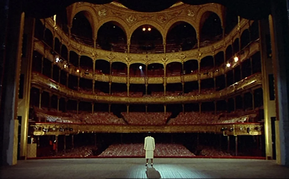 Diva (1981) movie still: wide — An opulent, multi-tiered historic theater with gold filigree and plush red seating stands empty. A lone woman in a…