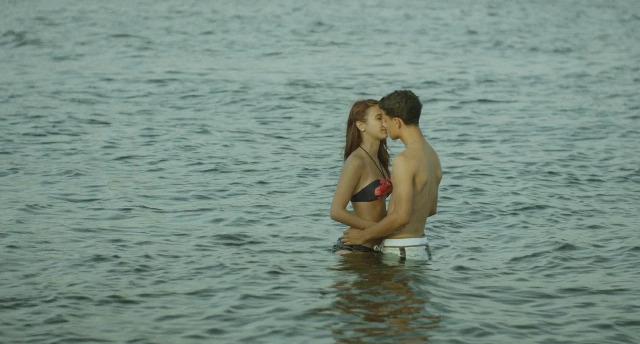 It Felt Like Love (2013) movie still: fullbody — A man and woman stand in calm, waist-deep water under natural light. The couple embraces and kisses…