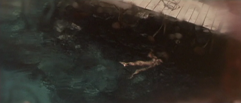 Boom! (1968) movie still: wide — A dark body of water sits adjacent to a weathered wooden plank structure. Riptides and surface textures are visible…