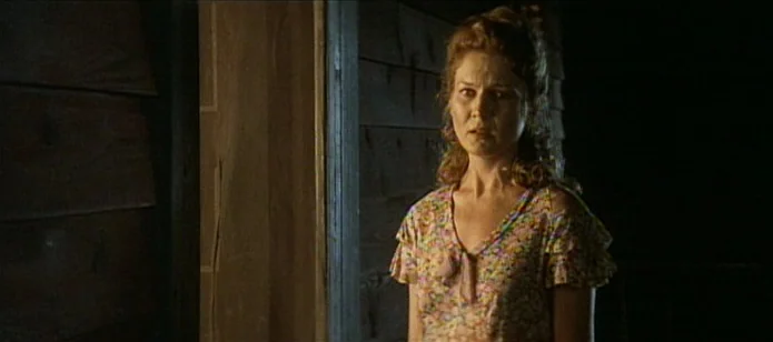 The Neon Bible (1995) movie still: medium — A woman stands in a doorway of a rustic wooden structure at night. She looks toward the side with a…