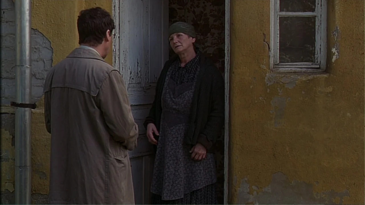 Citizen X (1995) movie still: medium — A man in a trench coat faces an older woman in a doorway. The exterior wall is yellow plaster with visible…
