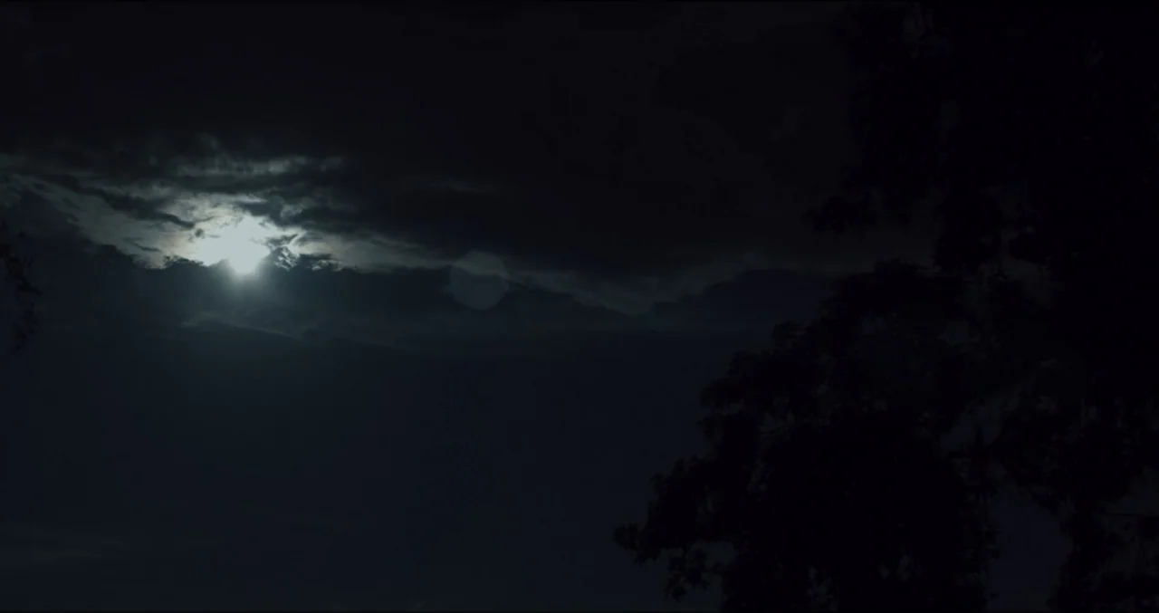 Eye for an Eye (2025) movie still: establishing — Dark storm clouds move across the night sky in front of a bright moon. The silhouette of tree…