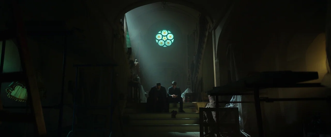 The Pope’s Exorcist (2023) movie still: wide — A dark, cluttered interior of an old church with draped furniture and stone stairs. Two men sit…