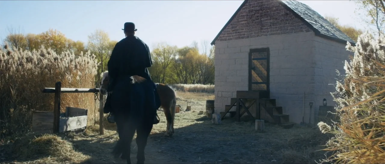 Out of Liberty (2019) movie still: medium — A rider on a brown horse approaches a small, single-story stone structure with brick accents. The…
