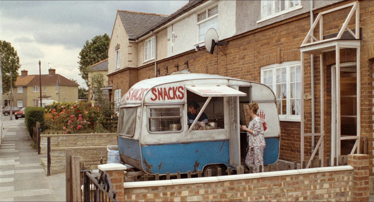 Life Is Sweet (1990) movie still: wide — A residential suburban street with rows of brick houses and a snack trailer parked. A man prepares food…
