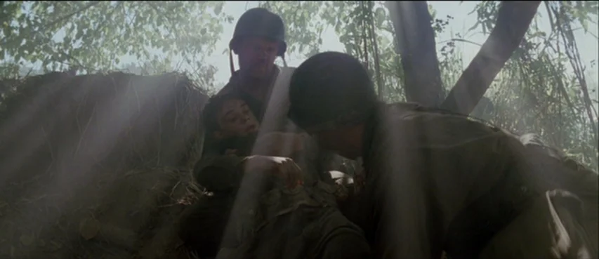 The Thin Red Line movie still: medium — Three soldiers crouch among dirt and leaves in a dense wooded area. Two soldiers attend to a third soldier…