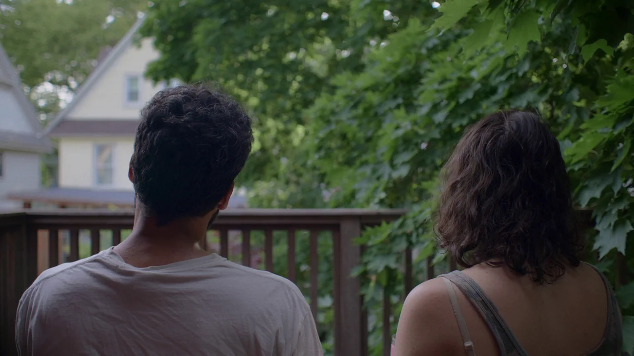 The Feeling That the Time for Doing Something Has Passed (2023) movie still: medium — An outdoor balcony overlooking a green leafy tree and a…