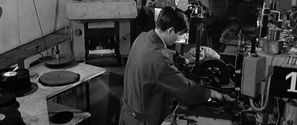 Antoine and Colette (1962) movie still: medium — An industrial factory floor contains heavy mechanical equipment used for pressing vinyl records. A…