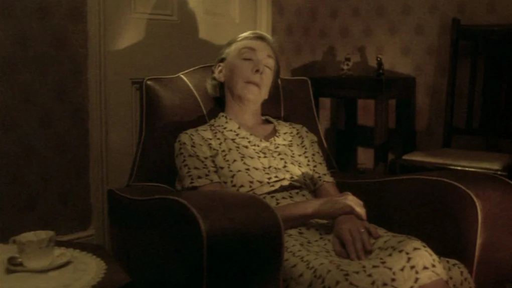 Distant Voices, Still Lives (1988) movie still: medium — A dimly lit room features a patterned armchair occupied by an elderly woman. The subject…