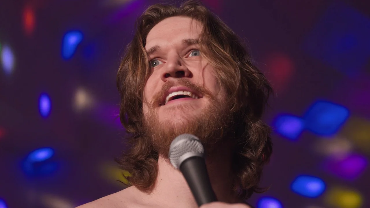 Bo Burnham: Inside (2021) movie still: closeup — A man stands on a stage with a background of out-of-focus colorful lights. He holds a microphone…
