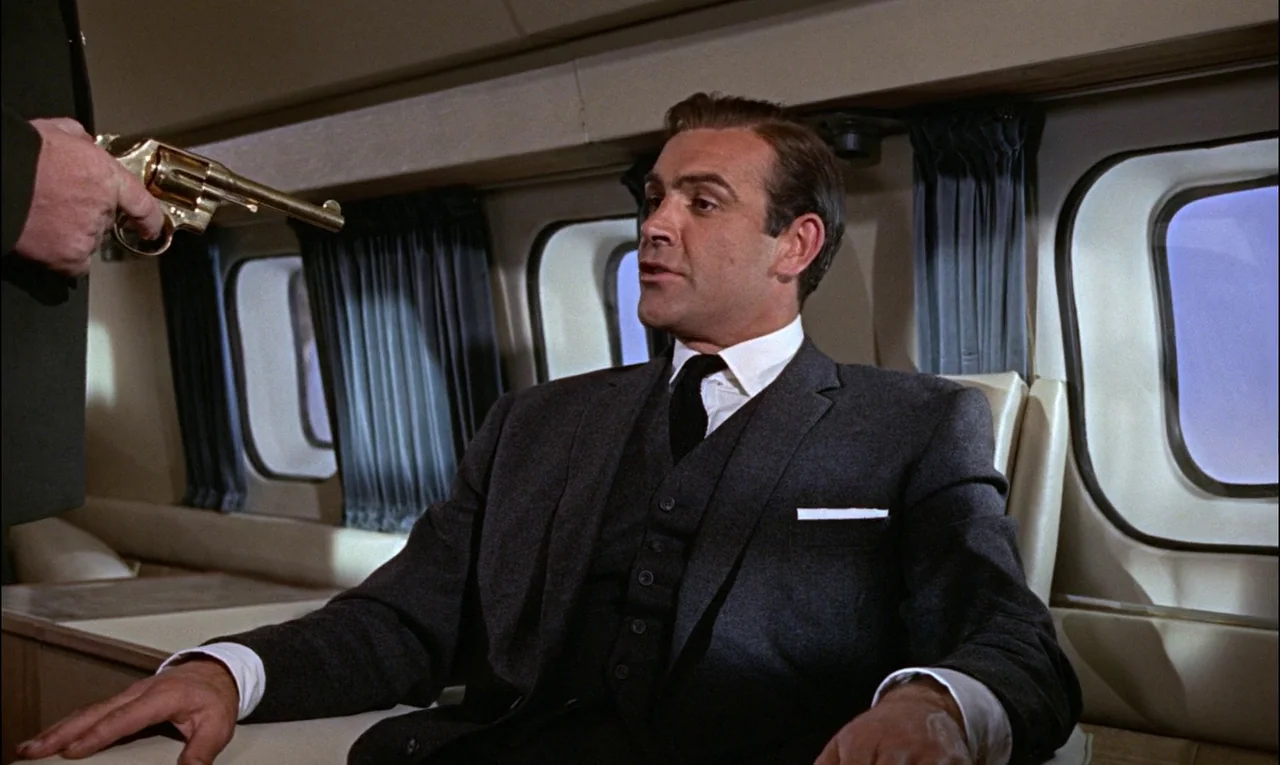 Goldfinger (1964) movie still: medium — The interior of an airplane cabin features light-colored walls and curtains by the windows. A man in a…