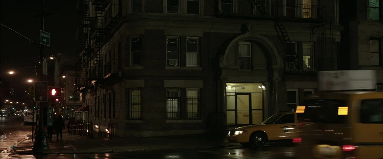 Collateral Beauty (2016) movie still: wide — A dark brick apartment building sits on a city street corner at night. A yellow taxi speeds past the…