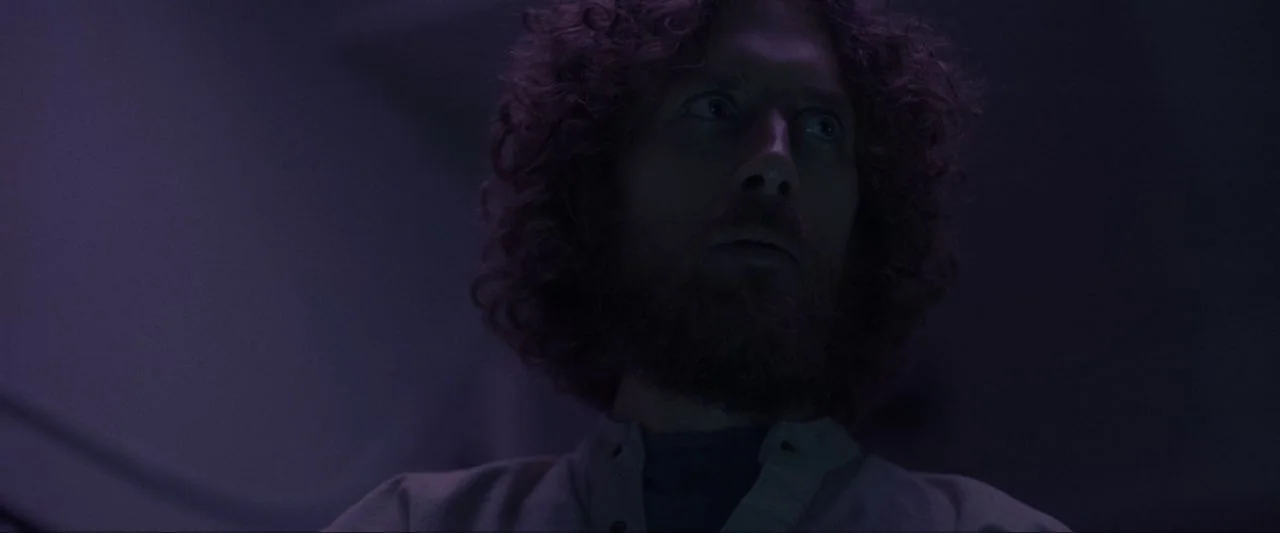 Come True (2020) movie still: closeup — A dimly lit indoor space is bathed in monochromatic purple light. A man with curly hair and a beard stares…
