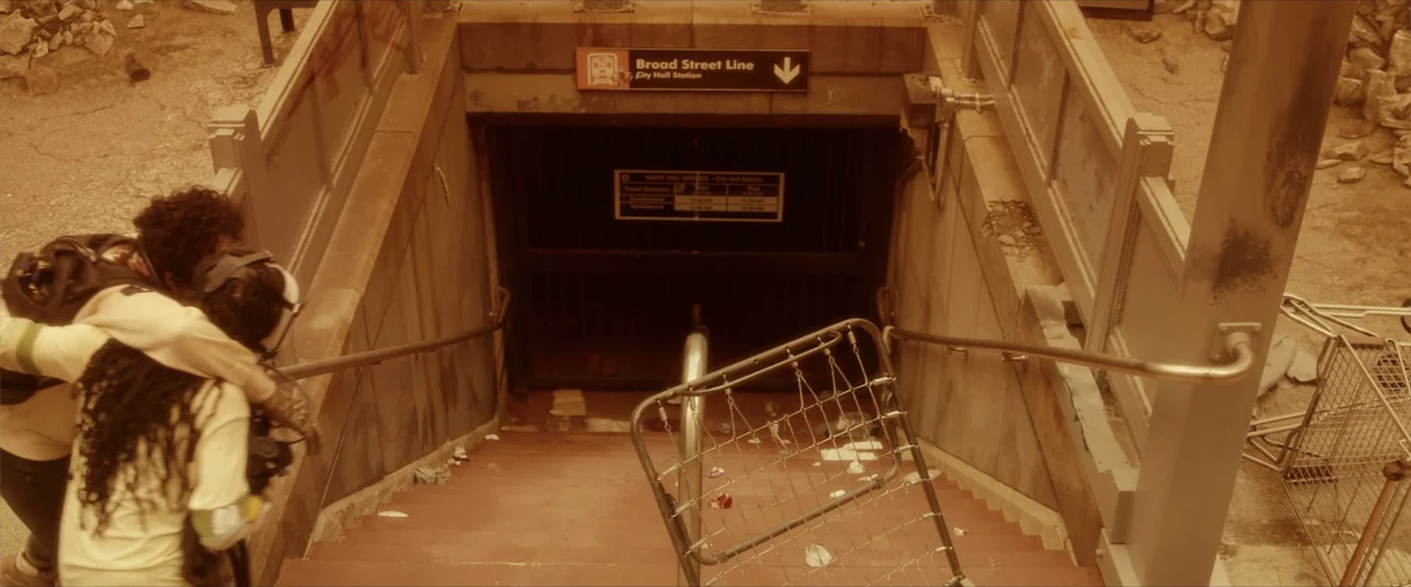 Breathe (2011) movie still: medium — A dirty subway station entrance with metal railings and scattered debris on the steps. One individual carries…