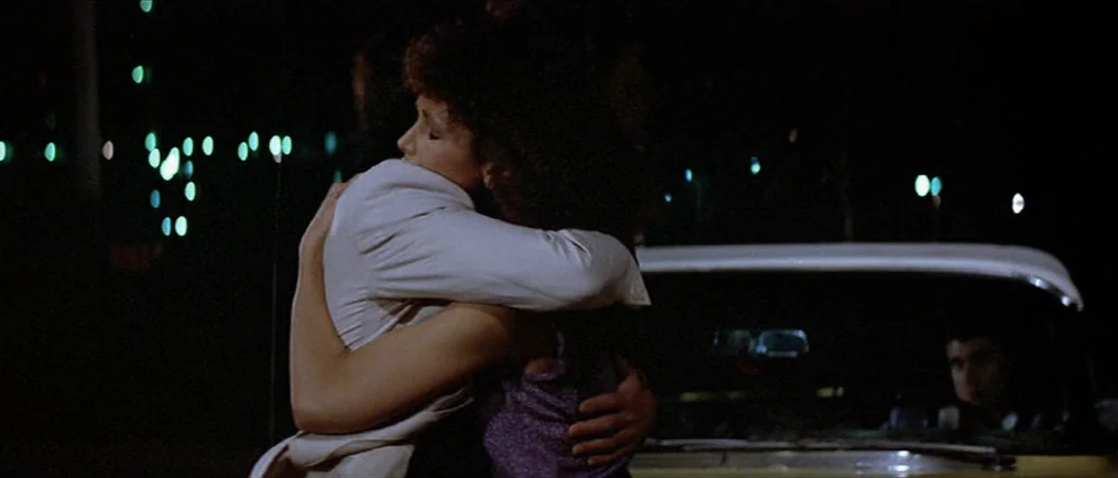 Scarface (1983) movie still: medium — A night scene in a dark parking lot with distant bokeh city lights. Two people are locked in a close embrace…