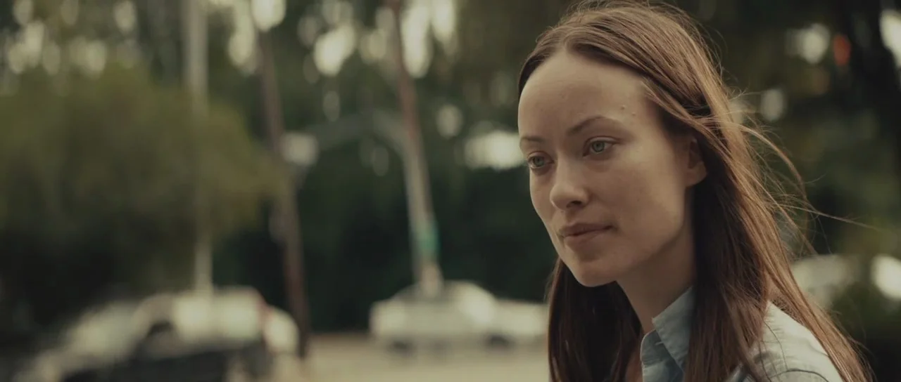 Meadowland (2015) movie still: closeup — A woman stands outdoors in an urban setting with blurred cars and trees behind her. She looks toward the…