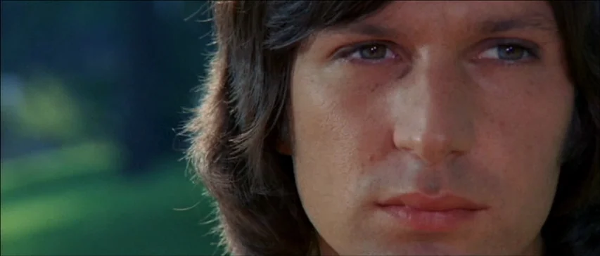 Four Flies On Grey Velvet (1971) movie still: closeup — A close-up of a young man against a soft-focus, verdant outdoor background. The subject…
