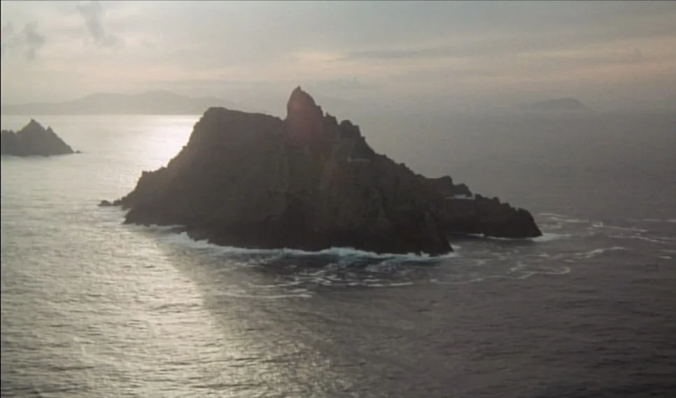 Heart of Glass (1976) movie still: establishing — A rocky island rises from the ocean under a hazy sunset sky. Dark craggy cliffs are surrounded by…