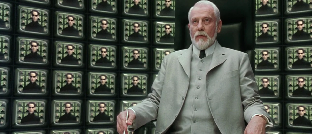 The Matrix Reloaded (2003) movie still: medium — A man sits in an office chair against a wall of many television monitors. Every screen displays the…