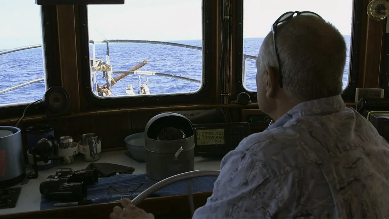 Zombie Sharks (2014) movie still: medium — The interior of a wooden boat cabin overlooks open blue ocean water through windows. A man sits at the…