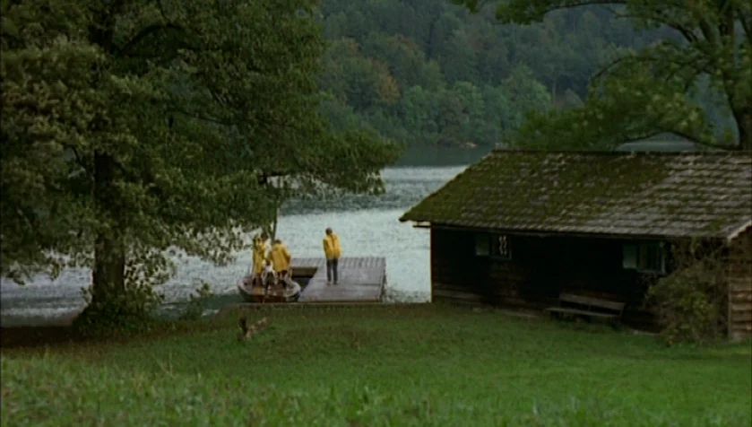 Funny Games (1997) movie still: wide — A grassy lakeside clearing features a wooden boat dock and a small cabin. Several figures in yellow raincoats…