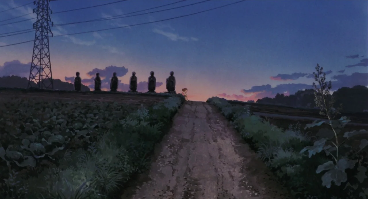 My Neighbour Totoro (1988) movie still: wide — A rural dirt path winds through fields under a deep blue twilight sky. Six small stone statues stand…