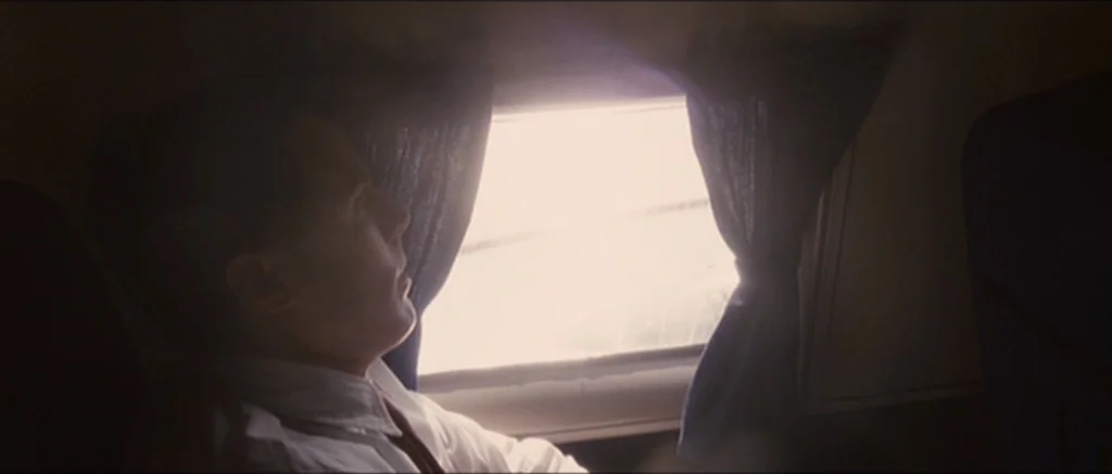 The Killer Inside Me (2010) movie still: medium — The interior of an airplane cabin features dark blue curtains flanking a bright window. A man in a…