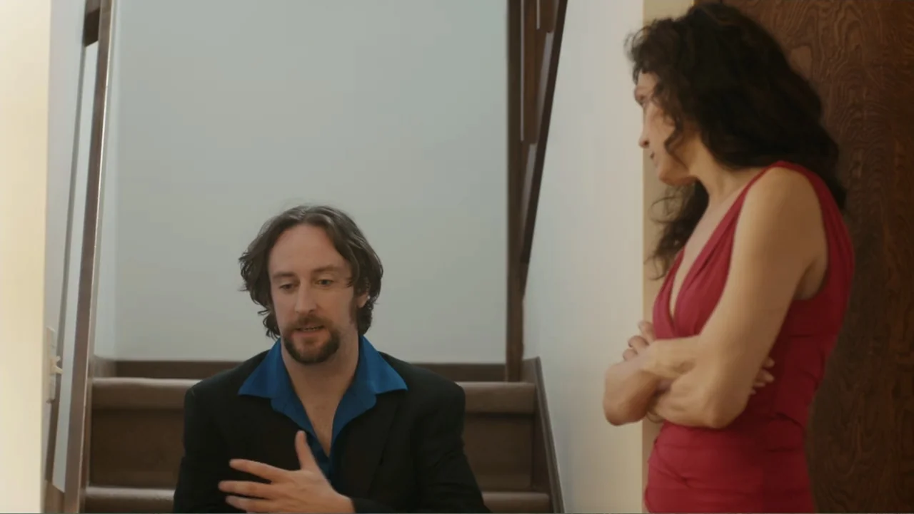 Toss It (2019) movie still: medium — A residential staircase with beige carpeting and a dark wooden door. A man talks to a woman as they stand on…