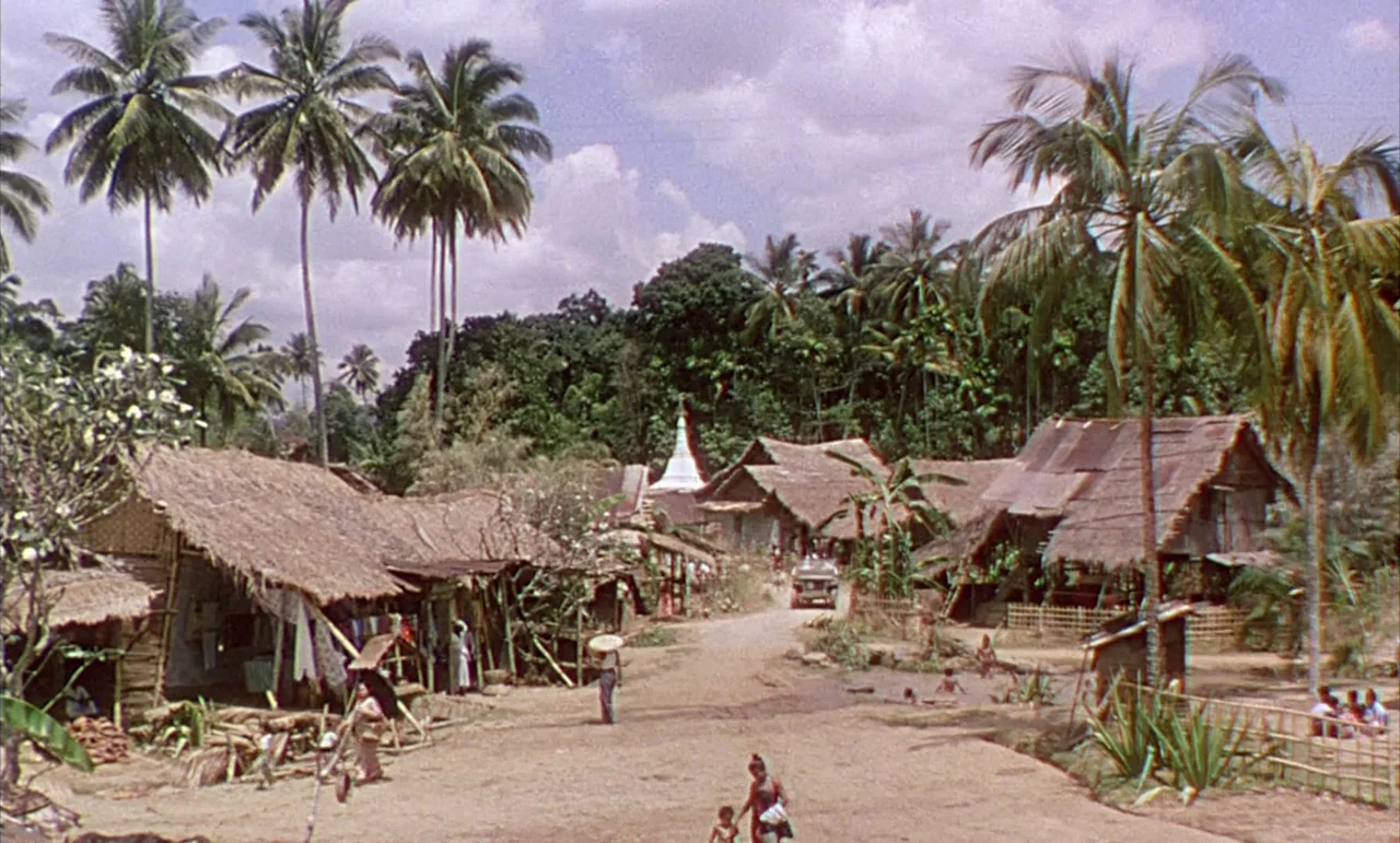 The Purple Plain (1954) movie still: establishing — A rural village with numerous thatched-roof wooden huts nestled among dense palm trees.…