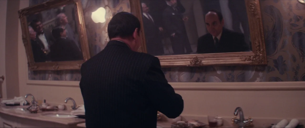 Neruda (2016) movie still: medium — A dimly lit upscale restroom features ornate gold mirrors and patterned wallpaper. A man in a suit looks at his…