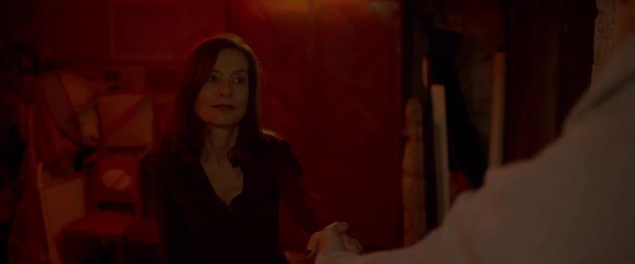 Elle (2016) movie still: medium — A dark, cluttered space is illuminated by a heavy, saturated red light. A woman in a dark shirt shakes the hand of…