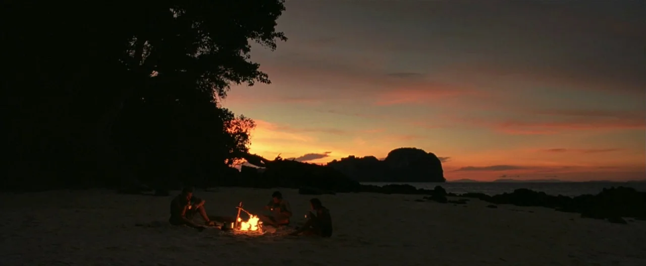 The Beach (2000) movie still: wide — A dark tropical beach shoreline under a dim orange and purple dusk sky. Three people sit around a small…