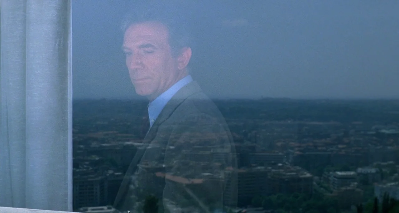 Tenebre (1982) movie still: medium — A man stands inside a room looking through a large glass window. The reflection of the urban city landscape…