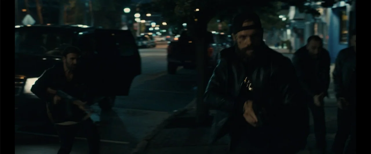 Howl (2015) movie still: medium — An urban street at night with a parked SUV in the background. A bearded man in a leather jacket holds a handgun…