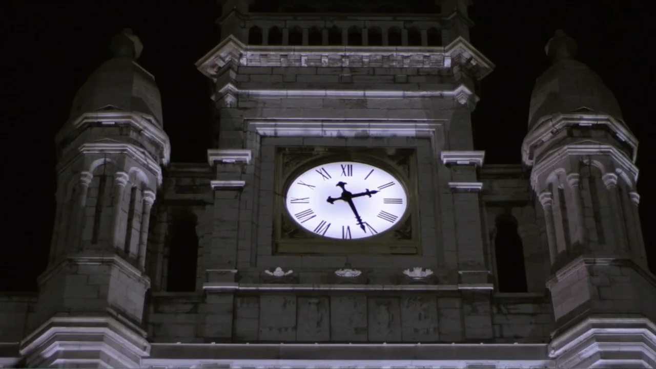 Entre las sombras (2025) movie still: wide — A dark stone clock tower rises against a black night sky. The illuminated clock face is centered with…