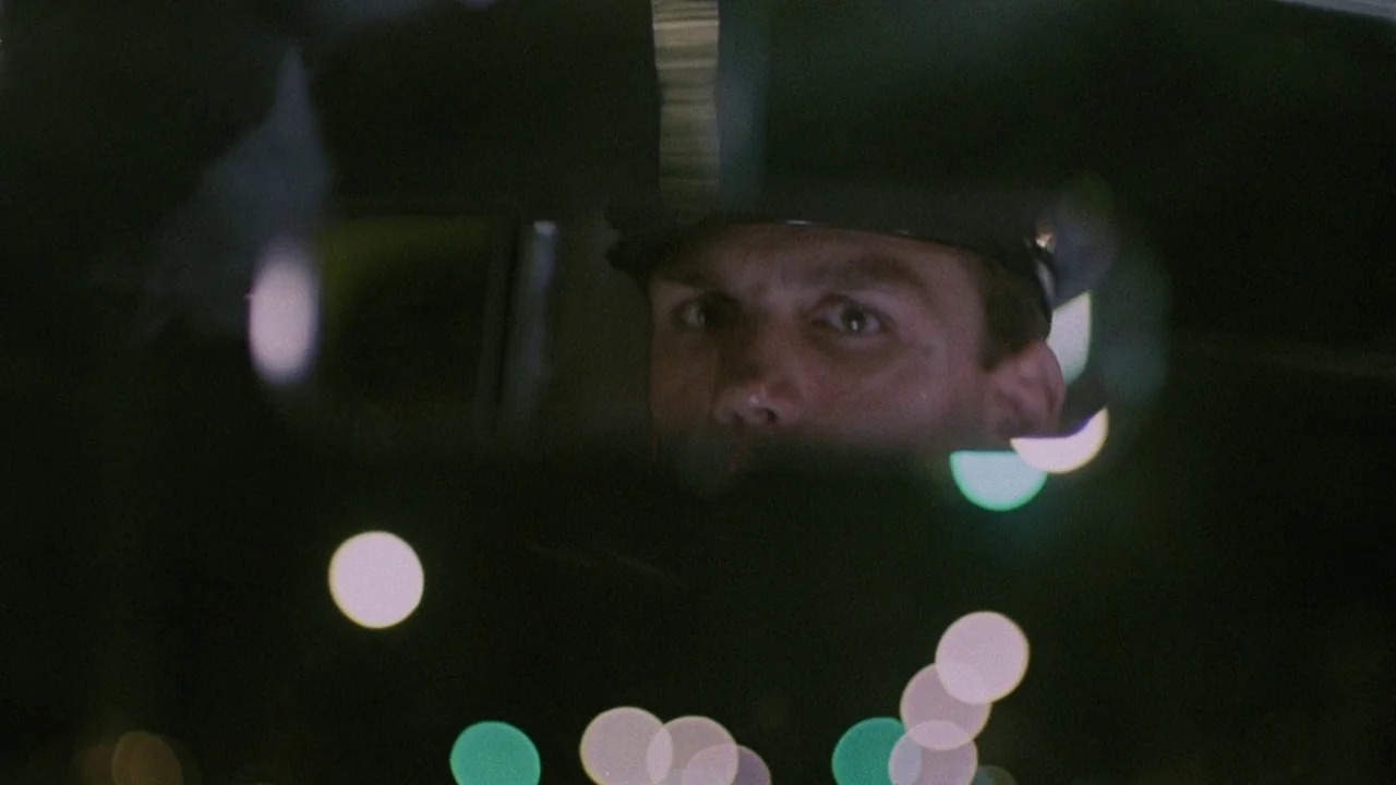 Insignificance (1985) movie still: closeup — The interior of a vehicle at night with a driver visible in the mirror. Bright out-of-focus city lights…